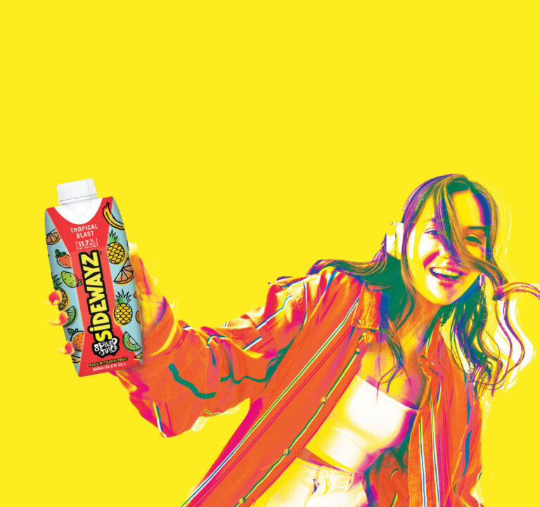 A smiling woman with long hair holds a colorful Sidewayz tropical fruit juice box against a bright yellow background. She wears headphones and a vibrant jacket, exuding energy and joy.