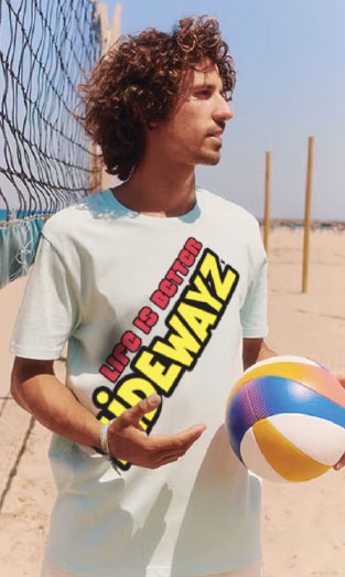 A man with curly hair stands on a sandy beach near a volleyball net, holding a colorful beach ball. He wears a T-shirt that reads LIFE IS BETTER SIDEWAYS in bold letters.