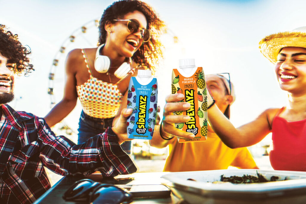 Four friends smile and toast with Sidewalkz juice cartons at an outdoor table. A Ferris wheel is visible in the background, suggesting a festival or fair setting under bright sunlight.