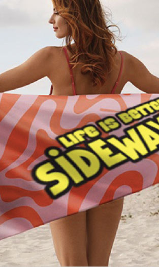 A woman in a pink bikini stands on a beach holding a colorful towel behind her that says Life is better sideways in bold letters.