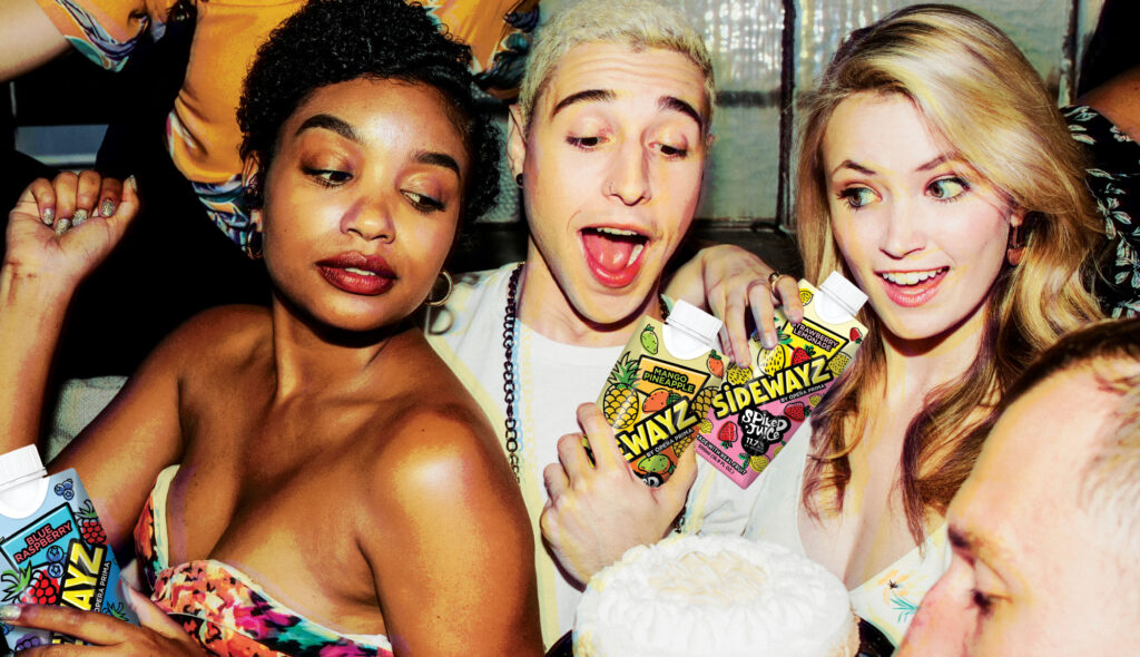 Three people at a lively party holding colorful Sidewayz juice cartons, smiling and posing together around a frosted cake, with bright, festive lighting and others in the background.