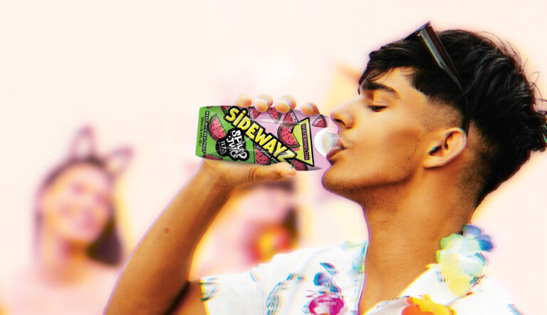 A young man wearing a floral shirt and lei drinks from a colorful can labeled SIDEWAYZ. The background is bright and blurred, with people and festive decorations visible.