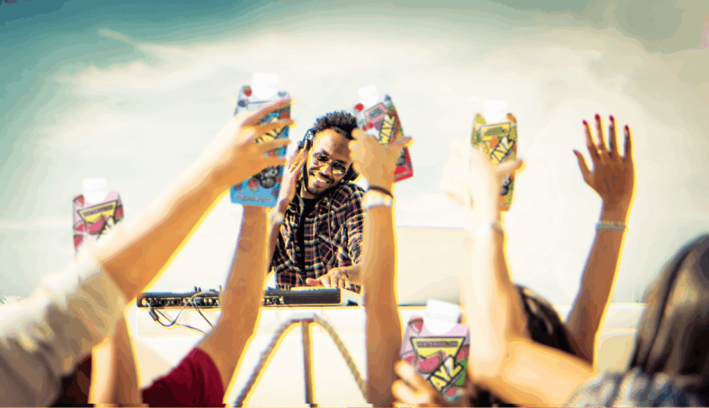 A DJ in sunglasses plays music outdoors as a crowd raises colorful drink cartons in the air, celebrating under a bright sky.