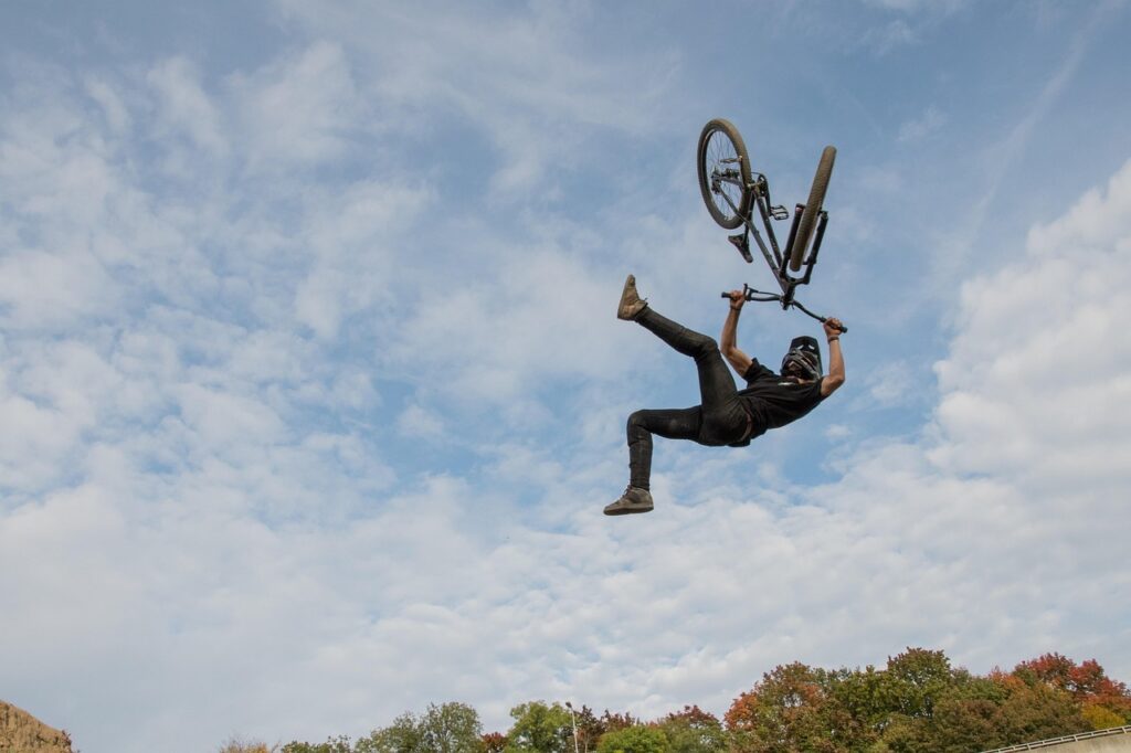 A person wearing a helmet performs an aerial trick on a BMX bike against a cloudy sky, with autumn trees and a hill in the background.