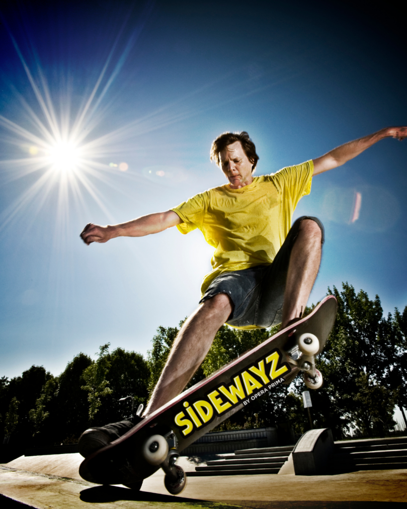 A person in a yellow shirt and shorts performs a skateboard trick at a skatepark under a bright sun, with SIDEWAYZ visible on the skateboard and trees in the background.