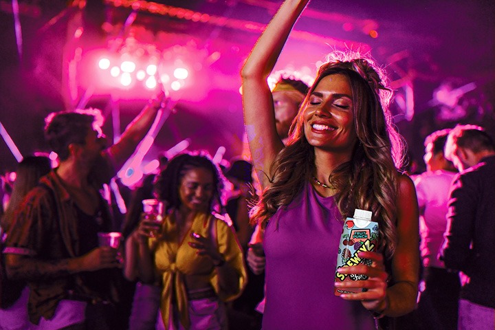 A woman in a purple top smiles with her eyes closed and raises one arm while holding a drink at a lively party, surrounded by people and colorful pink lights in the background.