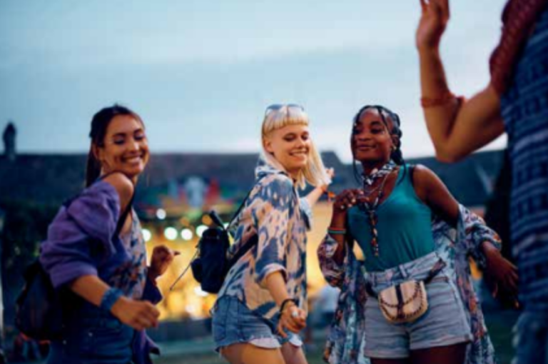 Three young women smile and dance outdoors at sunset, dressed in casual summer clothes. There are festival lights and blurred people in the background, creating a lively, joyful atmosphere.