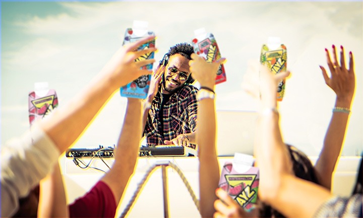 A DJ wearing sunglasses performs outdoors behind a turntable. People in the foreground raise their hands, holding up Sidewayz spiked juice, celebrating under a bright sky.